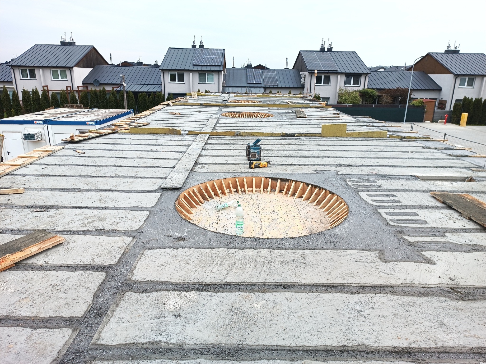 Betonowy strop w trakcie budowy z okrągłym otworem, na tle domów jednorodzinnych z szarymi dachami.