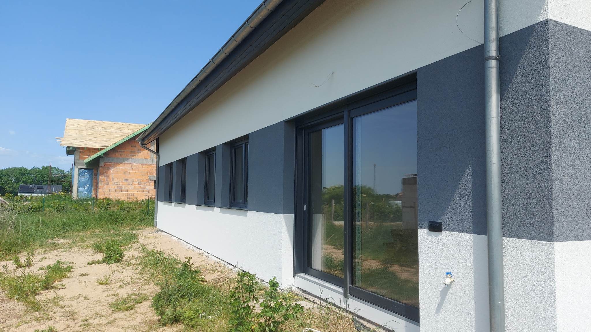Nowoczesny dom z elewacją w odcieniach szarości i bieli, widoczne okna z czarnymi ramami, obok budynek w trakcie budowy z odkrytą konstrukcją dachu.