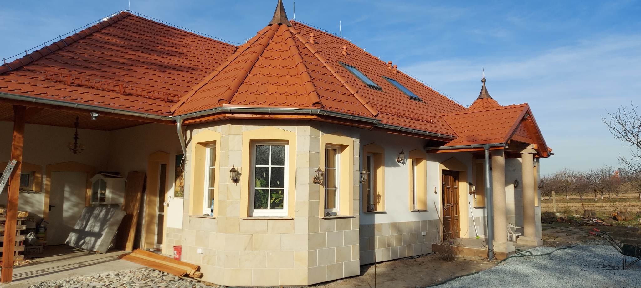 Widok z zewnątrz na dom z czerwoną dachówką, kamienną elewacją i oknami z białymi ramami w słoneczny dzień.