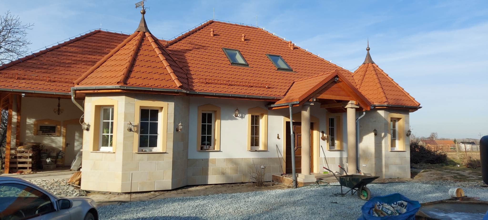 Dom jednorodzinny po tynkowaniu elewacji z elementami kamiennymi, widoczne detale architektoniczne, dachówka ceramiczna, teren wokół domu w trakcie zagospodarowania, taczka z narzędziami budowlanymi.