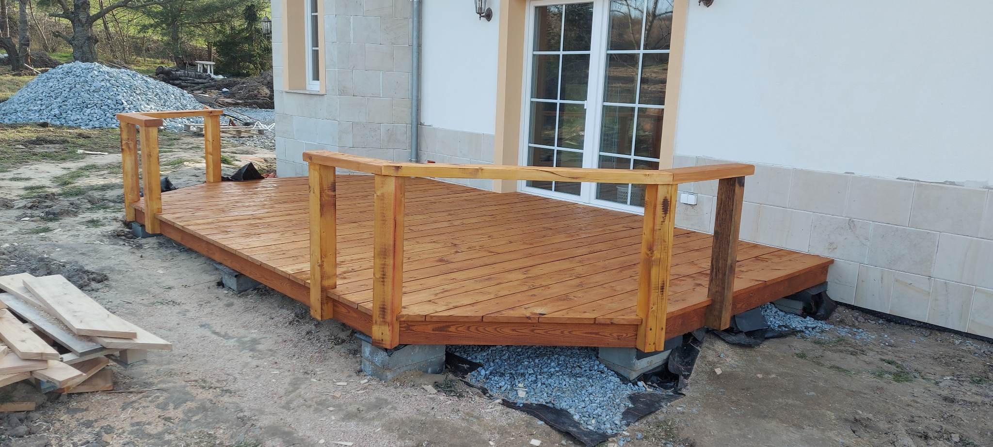 Nowo zbudowany taras z jasnego drewna z balustradą, oparty na betonowych blokach, z widoczną stertą gruzu i deskami w tle.