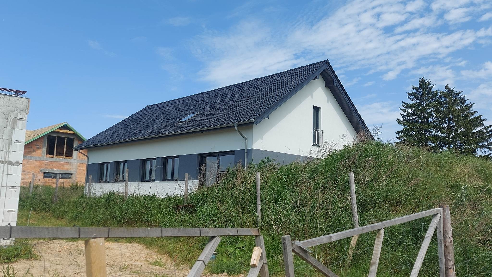 Nowoczesny dom jednorodzinny z dwukolorową elewacją (biało-szarą) i ciemną dachówką, widoczny zza wysokiej trawy i fragmentu drewnianego ogrodzenia, z budynkiem w trakcie budowy w tle po lewej...