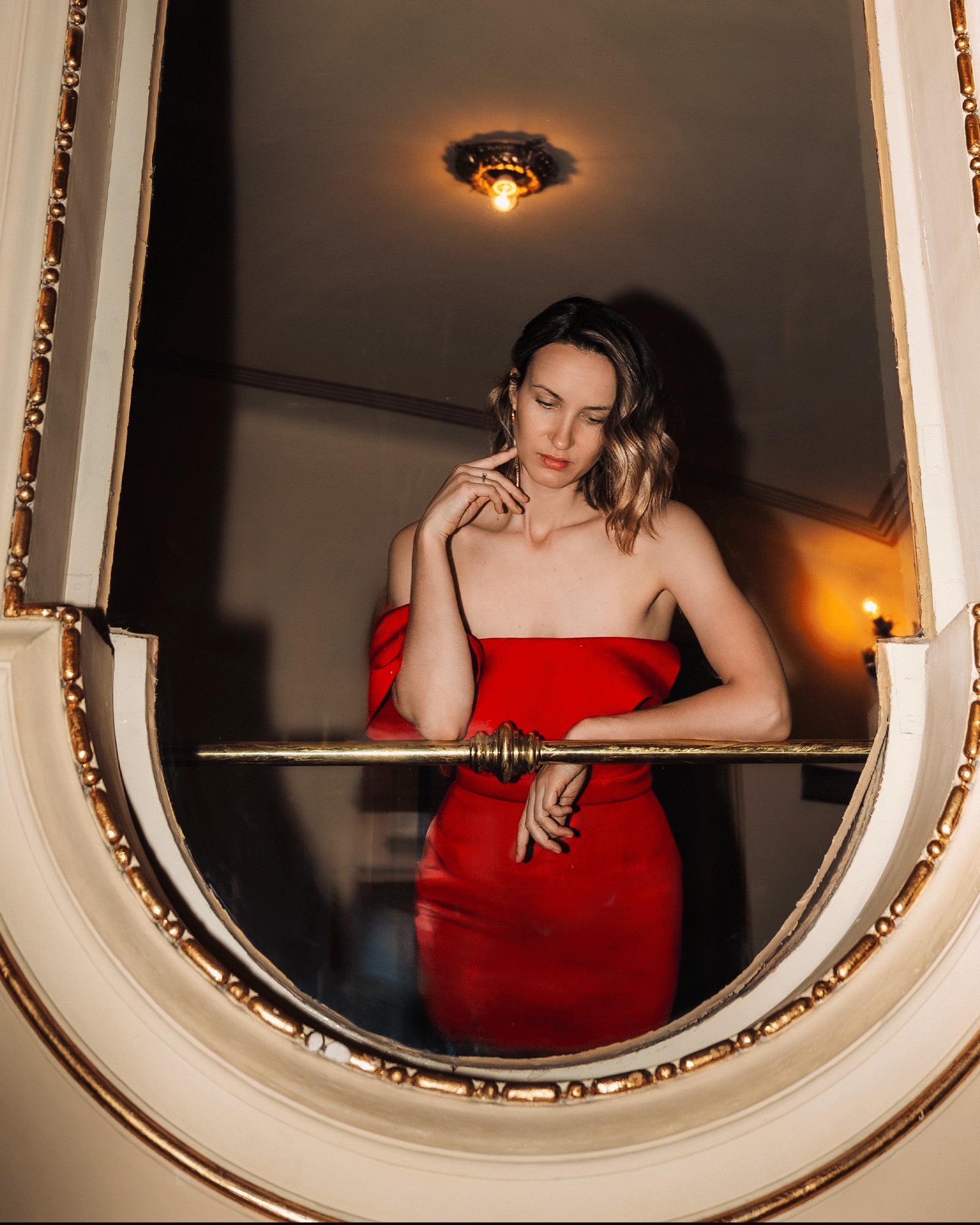Kobieta w czerwonej sukience, opierająca się o złotą balustradę, spoglądająca w dół, w ozdobnym, owalnym oknie.