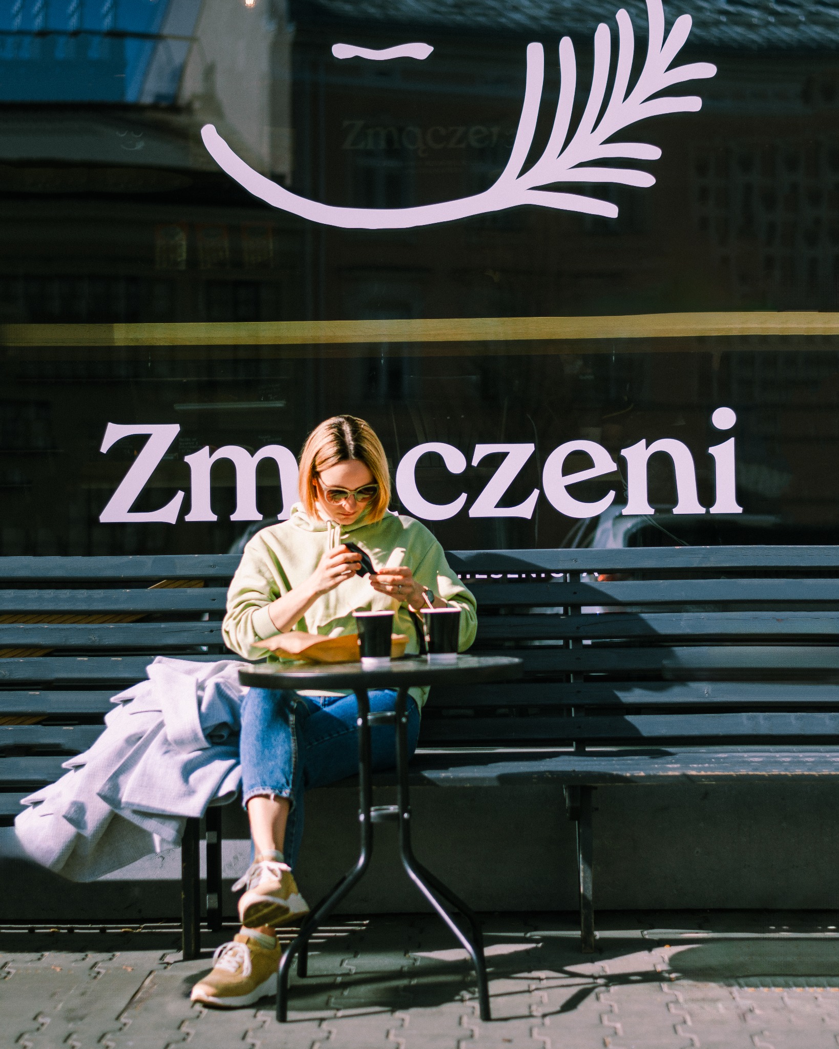 Kobieta w okularach siedzi przy stoliku przed kawiarnią Zmączeni, przeglądając telefon; na stoliku dwie kawy na wynos, na ławce leży szara kurtka.