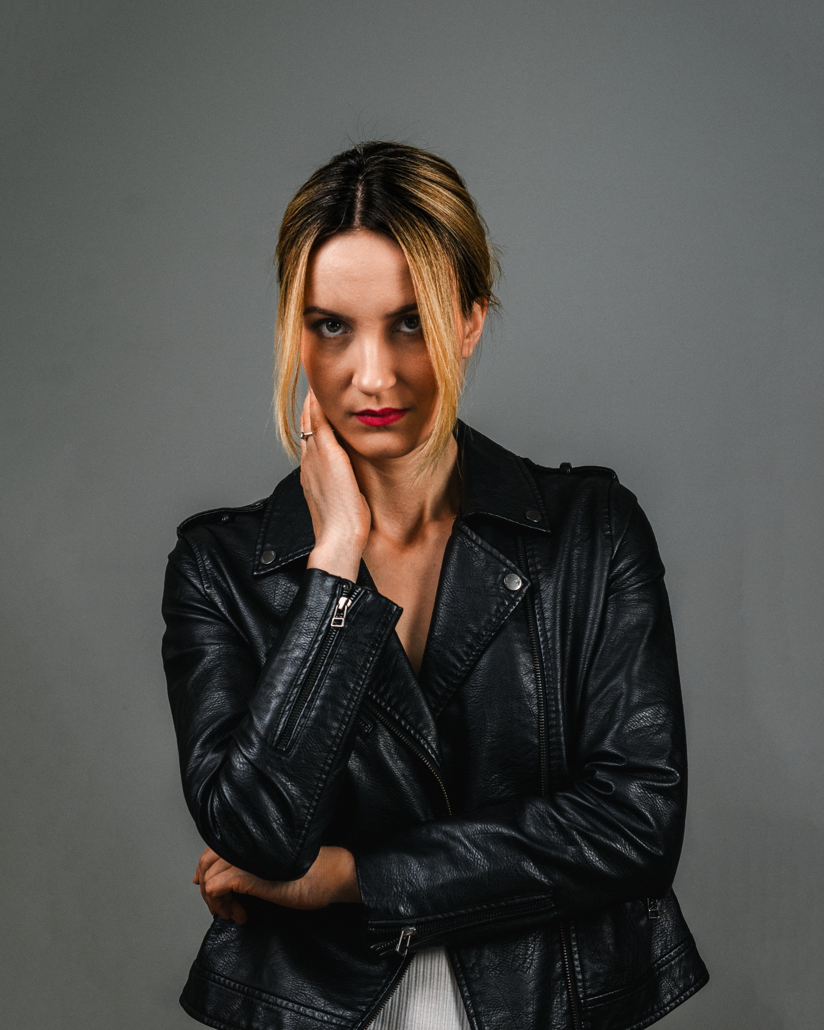 Portret kobiety w czarnej skórzanej kurtce na szarym tle, z dłonią przy twarzy i pierścionkiem na palcu.