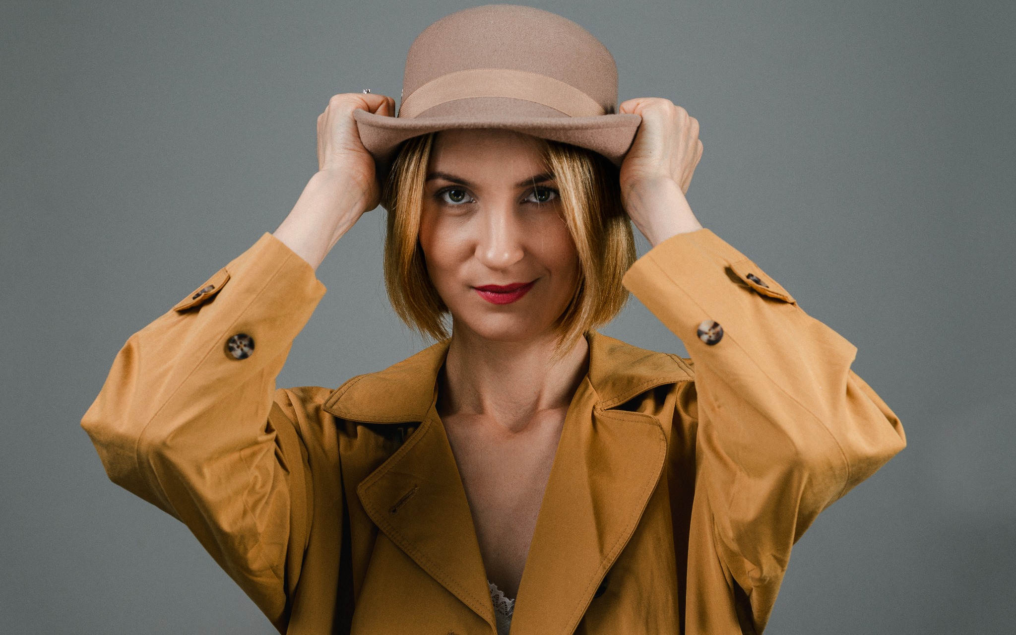 Portret kobiety w brązowym kapeluszu i płaszczu, z czerwonymi ustami, na szarym tle.