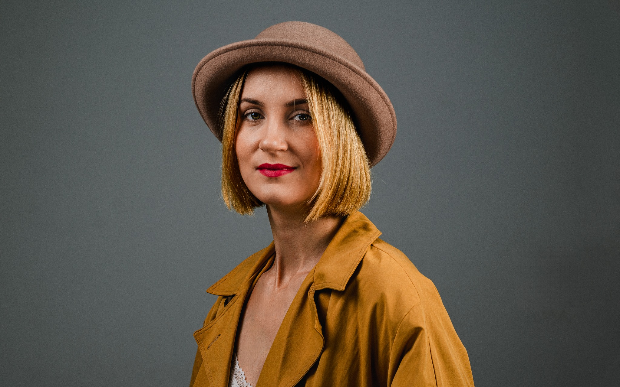 Portret kobiety w brązowym kapeluszu i musztardowym płaszczu, z czerwoną szminką, na szarym tle.