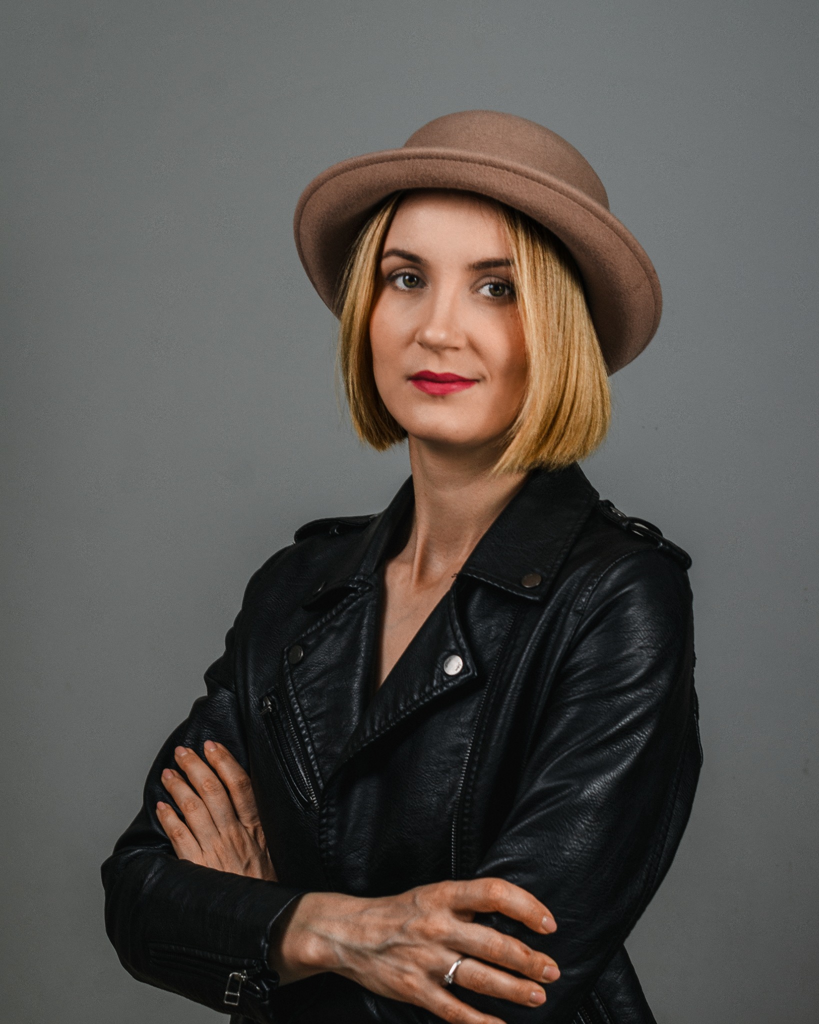 Portret kobiety w brązowym kapeluszu i czarnej skórzanej kurtce, z założonymi rękami, na szarym tle.