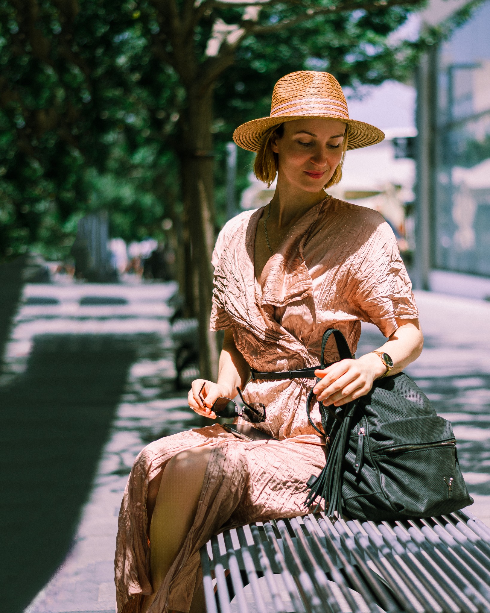 Kobieta w słomkowym kapeluszu siedzi na ławce w parku, trzymając okulary przeciwsłoneczne i czarny plecak.