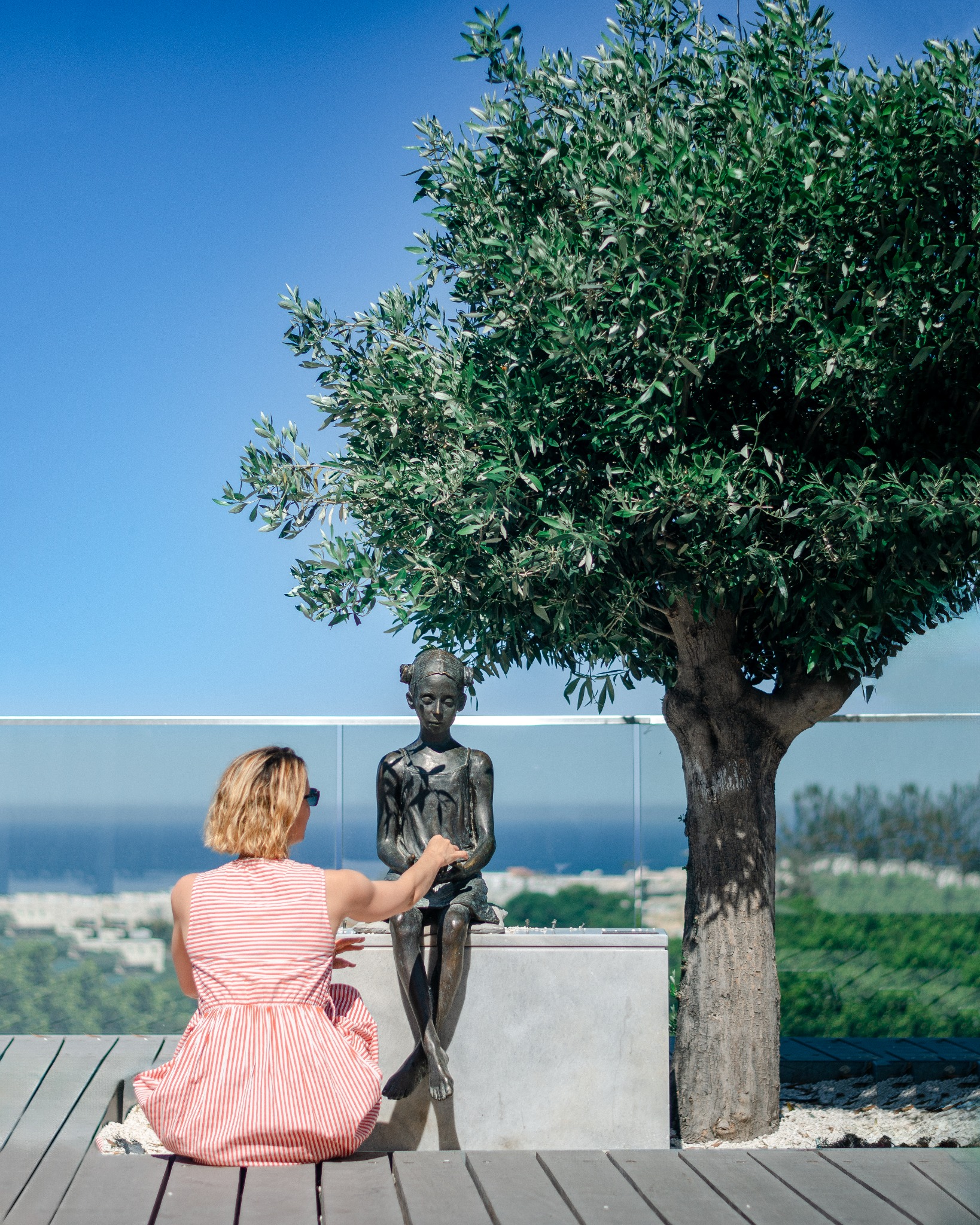 Kobieta w czerwono-białej sukience siedzi na drewnianej ławce obok posągu dziewczynki pod drzewem oliwnym, z widokiem na miasto i morze w tle.