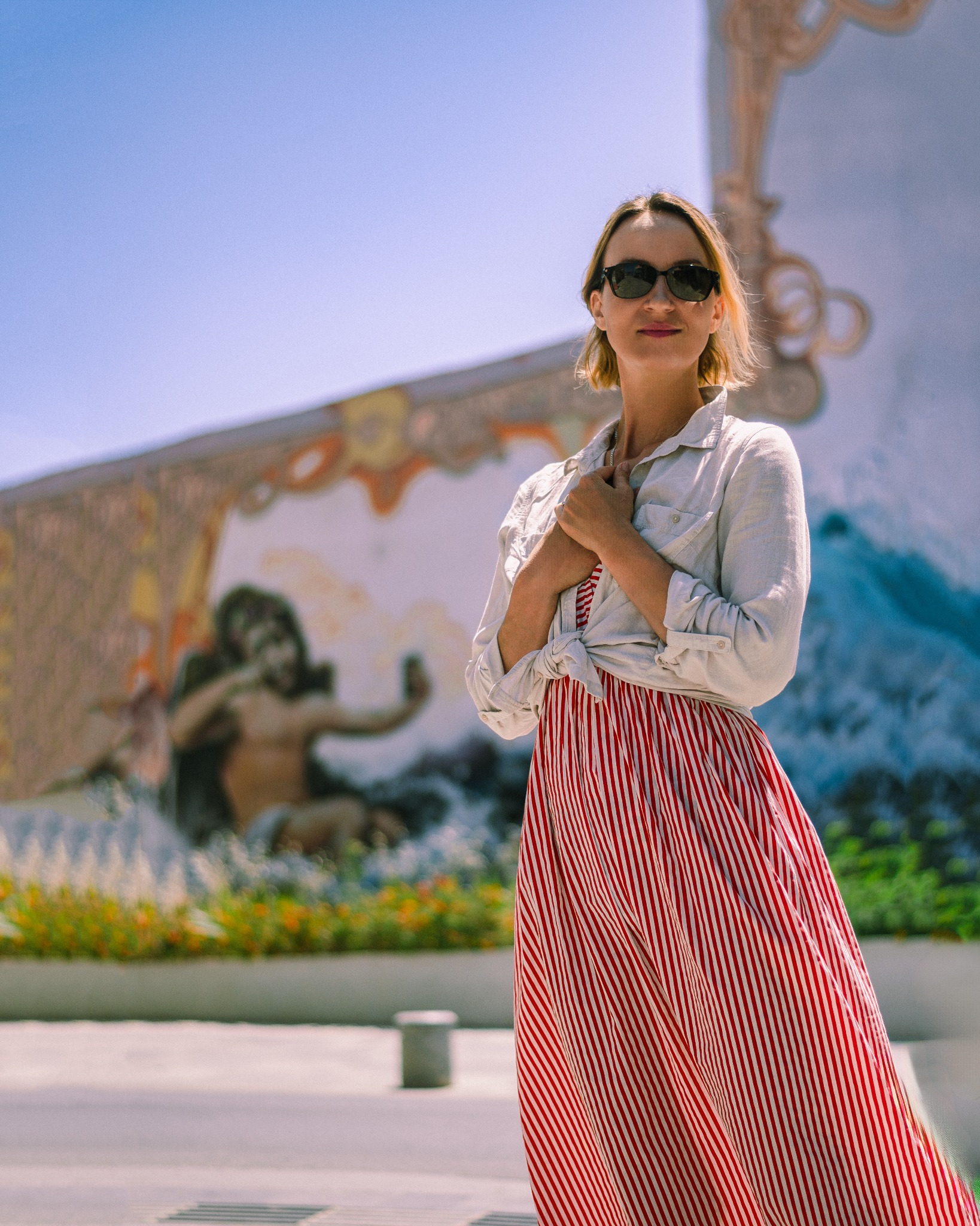 Kobieta w okularach przeciwsłonecznych, białej koszuli i czerwono-białej sukience w paski pozuje na tle kolorowego muralu z motywami mitologicznymi.