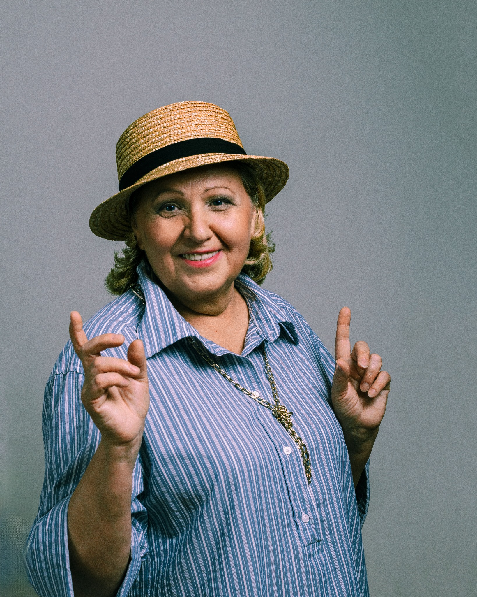 Uśmiechnięta kobieta w słomkowym kapeluszu i koszuli w paski, gestykulująca dłońmi na szarym tle.