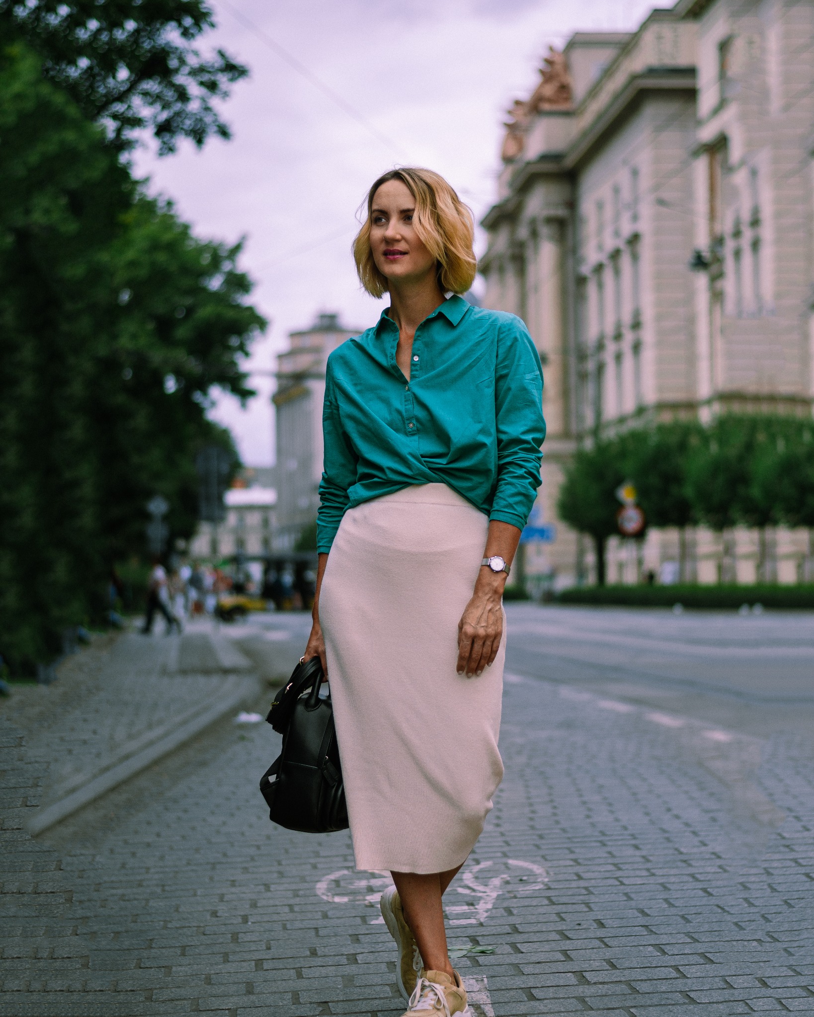 Kobieta w turkusowej koszuli i beżowej spódnicy pozuje na brukowanej ulicy z budynkiem w tle, trzymając czarny plecak.