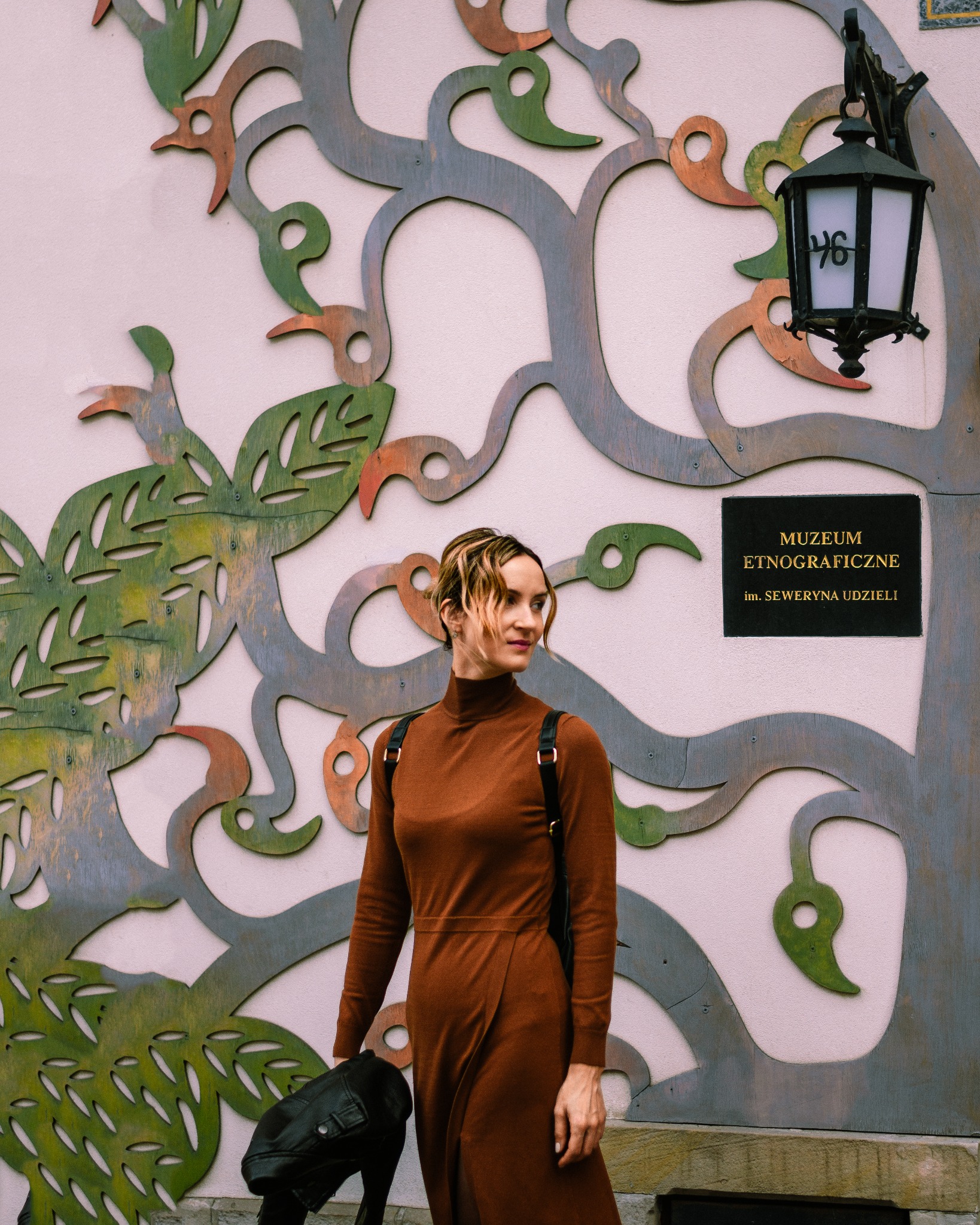 Kobieta w brązowej sukience stoi przed ścianą z dekoracją w kształcie drzewa i napisem Muzeum Etnograficzne im. Seweryna Udzieli.