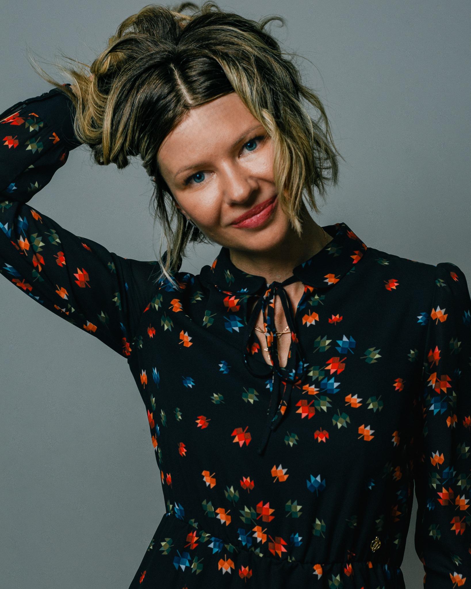 Portret uśmiechniętej kobiety z blond pasemkami, w czarnej sukience w kolorowe liście, pozującej na szarym tle z ręką we włosach.