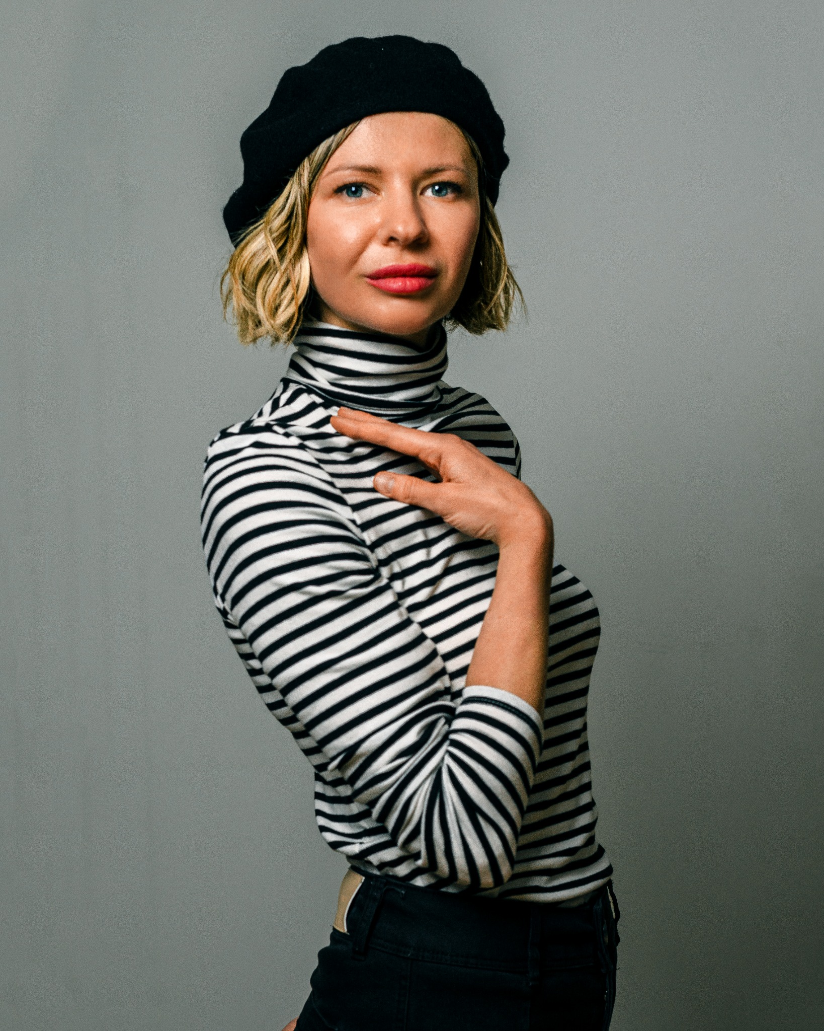 Portret kobiety w czarnym berecie i bluzce w czarno-białe paski, pozującej na szarym tle, z dłonią ułożoną na szyi.