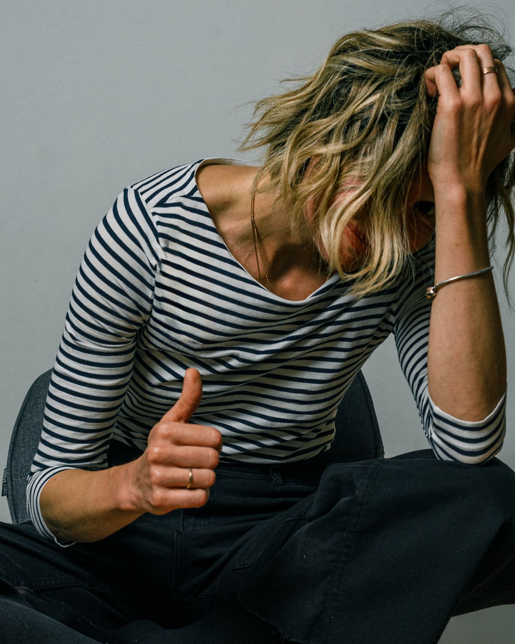 Kobieta w bluzce w paski siedzi na krześle, z jedną ręką na głowie i drugą podniesioną w geście kciuka w górę.