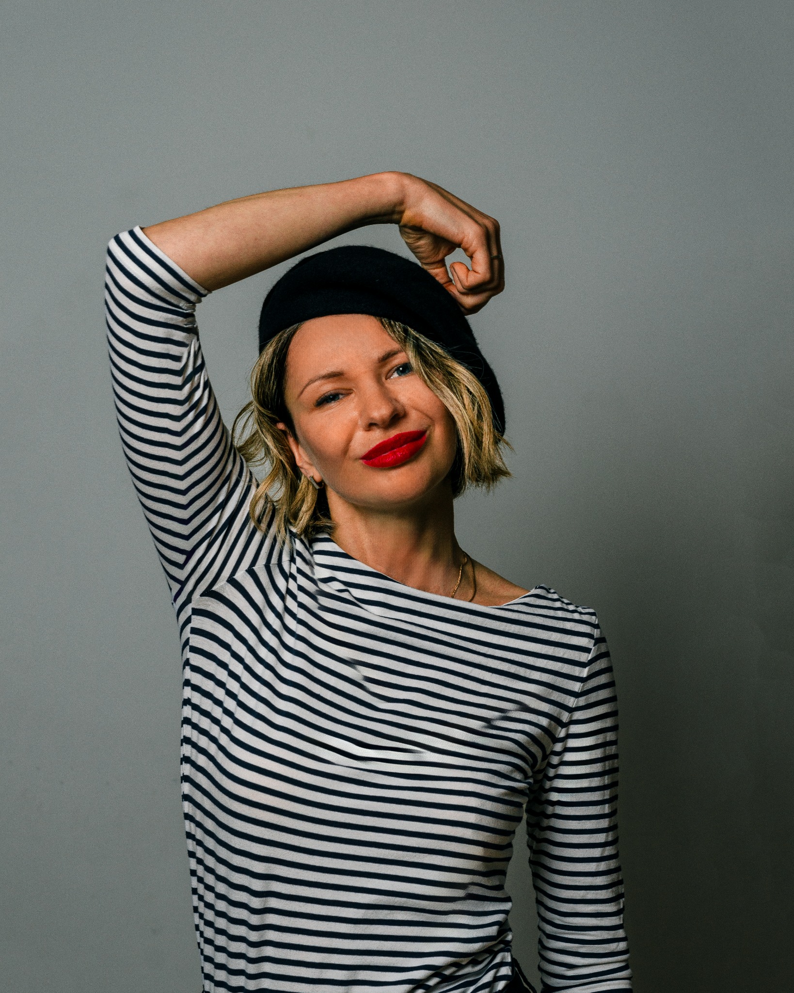 Portret kobiety w bretońskim topie i czarnym berecie, z ręką uniesioną nad głową, uśmiechającej się z czerwonymi ustami na szarym tle.