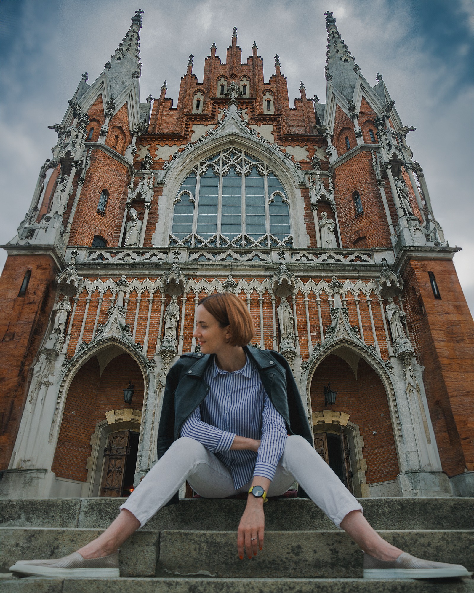 Kobieta w paski siedzi na schodach kościoła św. Józefa w Krakowie, ubrana w białe spodnie, skórzaną kurtkę i modny zegarek.