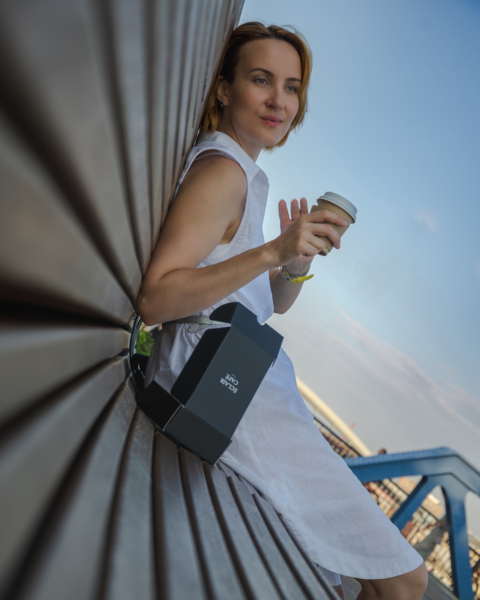 Kobieta w białej sukience siedzi na ławce, trzymając kubek z kawą i opierając się o czarne pudełko z napisem Eclair Cafe, z widokiem na błękitne niebo.