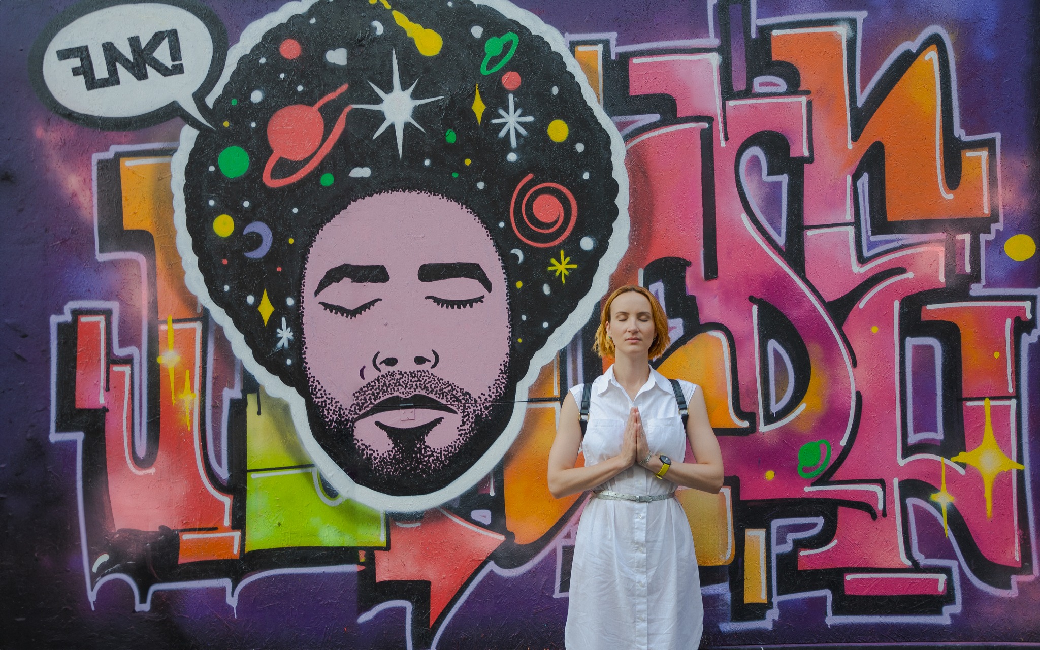 Kobieta w białej sukience stoi na tle kolorowego graffiti z wizerunkiem twarzy z fryzurą afro wypełnioną motywami kosmicznymi, ręce złożone w geście modlitwy.