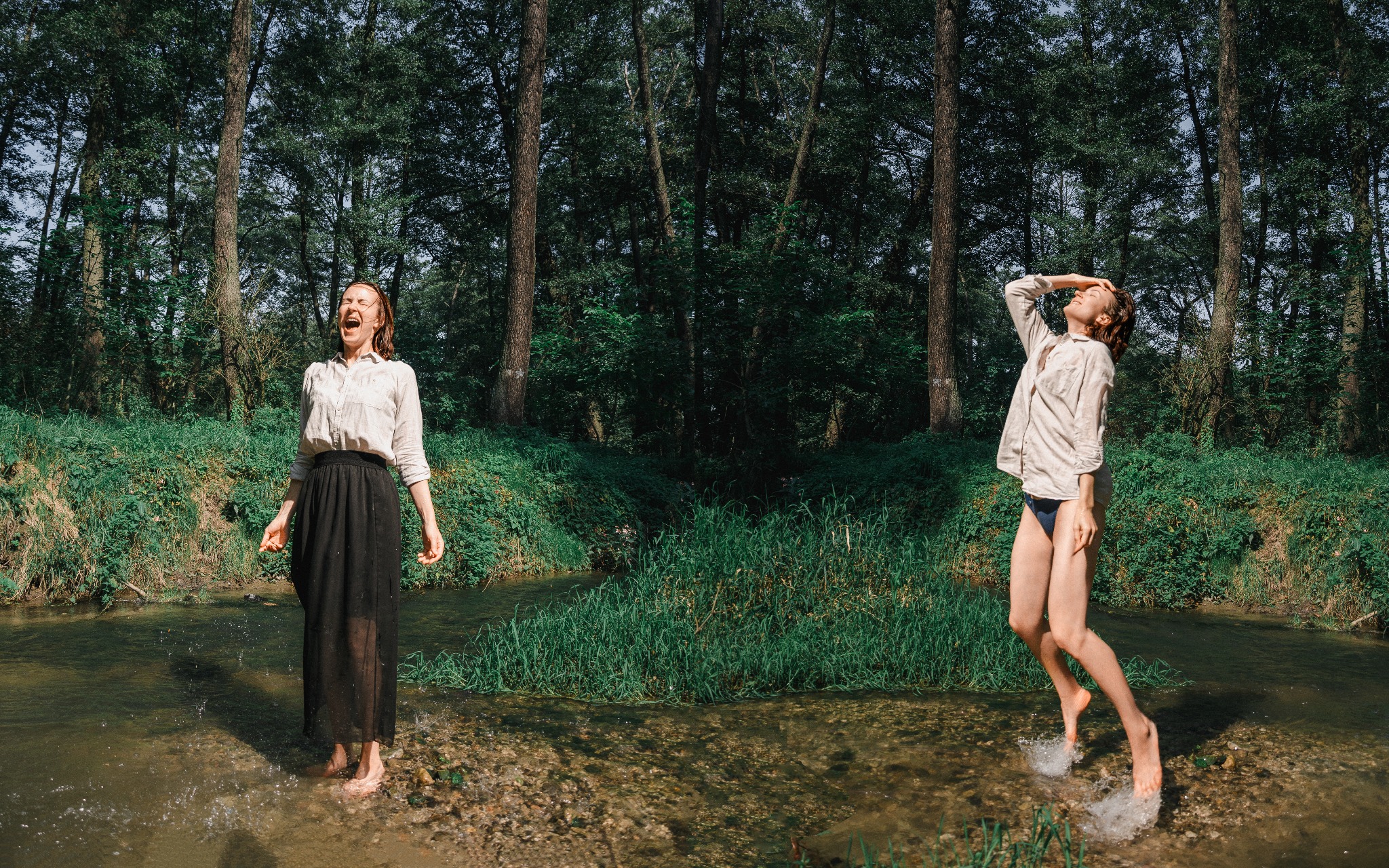 Dwie kobiety w rzece na tle lasu; jedna w długiej czarnej spódnicy i białej koszuli krzyczy, druga w bieliźnie i białej koszuli skacze, dotykając czoła.
