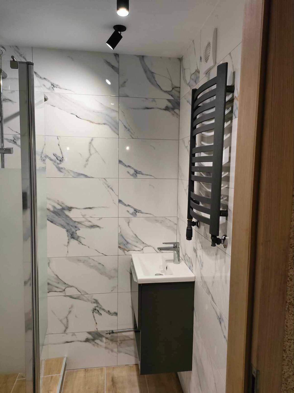 Nowoczesna łazienka z biało-szarymi płytkami imitującymi marmur, czarnym grzejnikiem drabinkowym i szafką pod umywalką w kolorze grafitowym, oświetlona punktowym światłem.