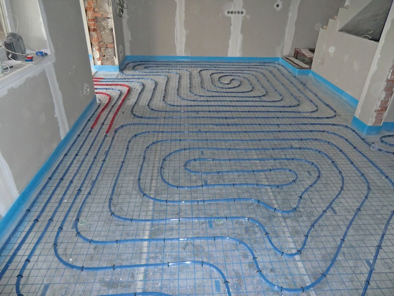Instalacja ogrzewania podłogowego w pomieszczeniu w stanie surowym, widoczne niebieskie rurki ułożone na metalowej siatce, ściany przygotowane do wykończenia.