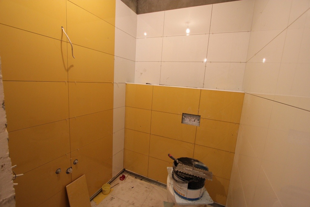 Żółte i białe płytki ceramiczne ułożone w łazience, widoczny kubeł z zaprawą i narzędzia glazurnicze, instalacja elektryczna przygotowana do podłączenia.
