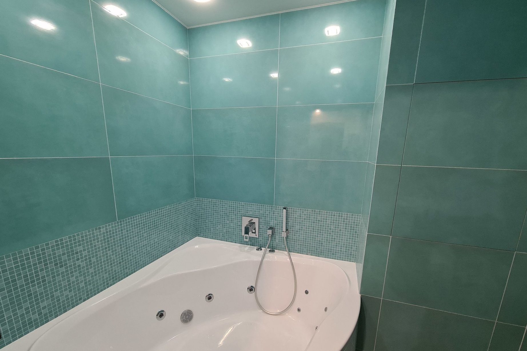 Turkusowa łazienka z białą wanną z hydromasażem, wykończona mozaiką w pasującym odcieniu, oświetlona punktowymi lampami sufitowymi.