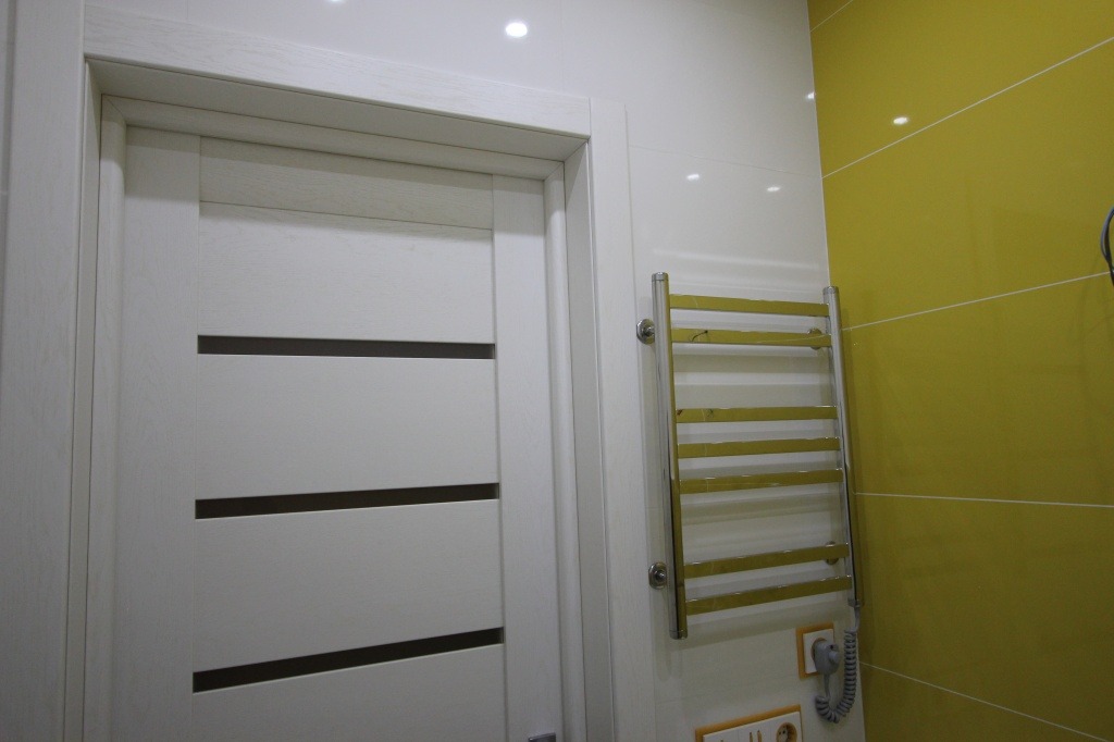 Białe drzwi wewnętrzne z poziomymi, ciemnymi intarsjami, zamontowane w łazience z grzejnikiem drabinkowym i żółtymi płytkami.