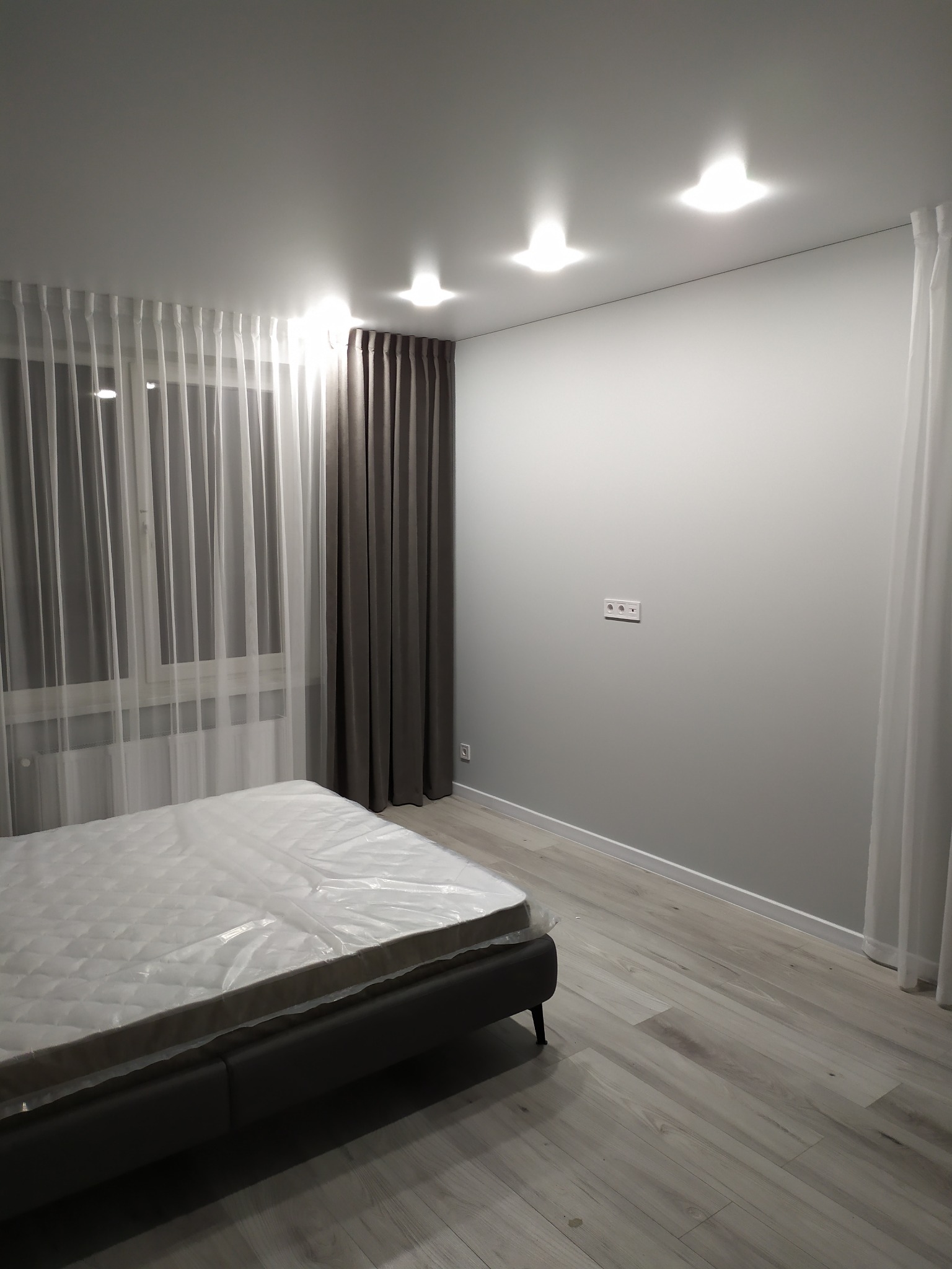 Pokój z łóżkiem w folii ochronnej, szare ściany, drewnopodobna podłoga, okno z białymi firankami i szarymi zasłonami, trzy punktowe lampy sufitowe.