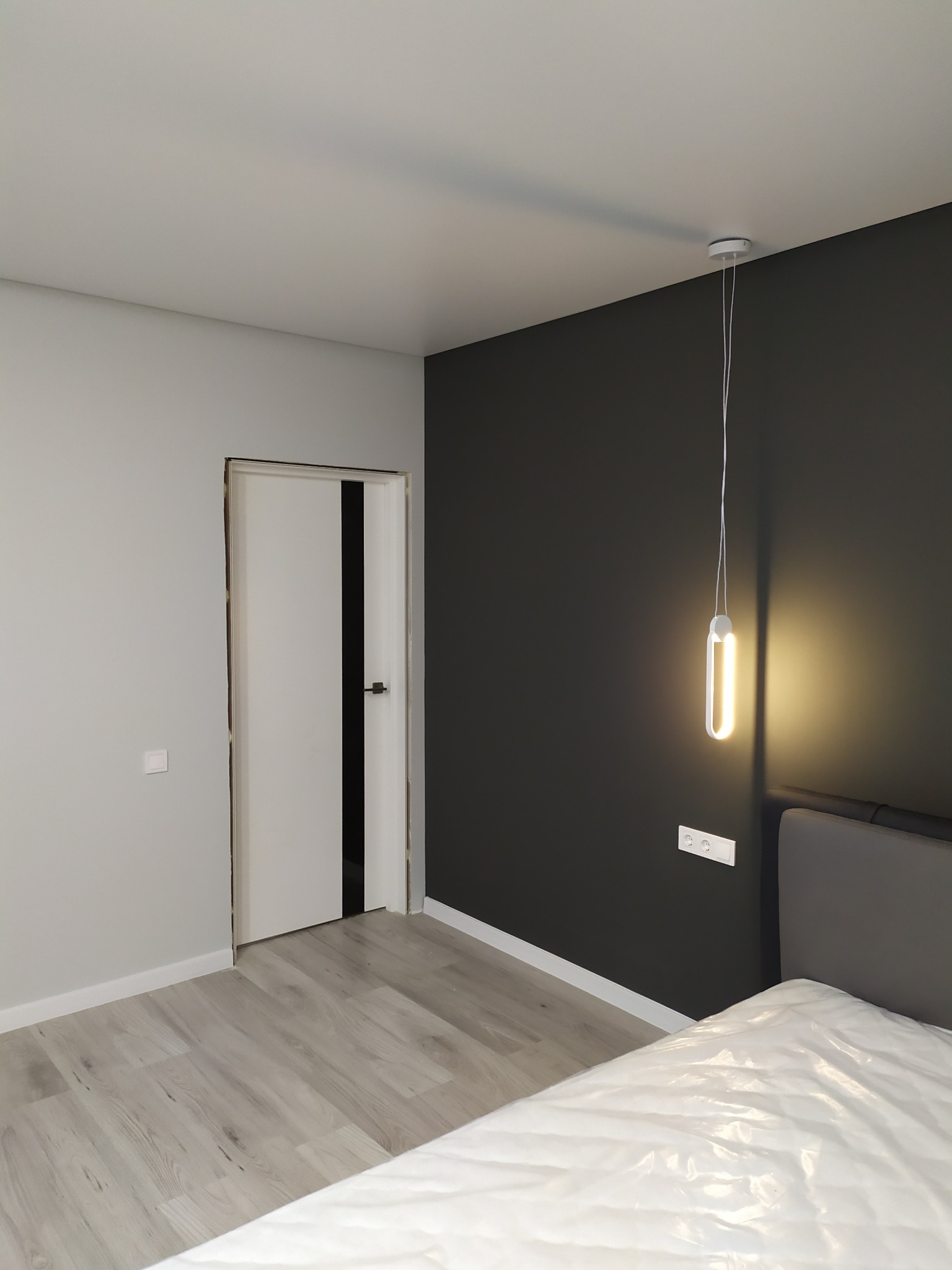 Nowoczesne wnętrze sypialni z szarą ścianą, białą lampą wiszącą o podłużnym kształcie i częściowo widocznym łóżkiem przykrytym folią ochronną; widoczny również fragment podłogi z panelami i białe...