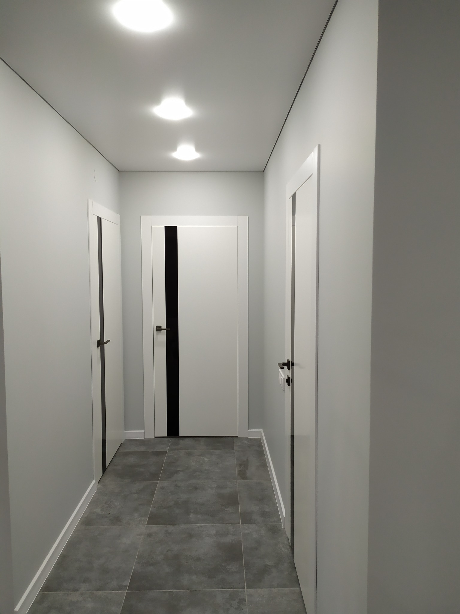 Korytarz w odcieniach szarości z trzema białymi drzwiami z czarnymi akcentami, szara podłoga z płytek, oświetlenie sufitowe punktowe
