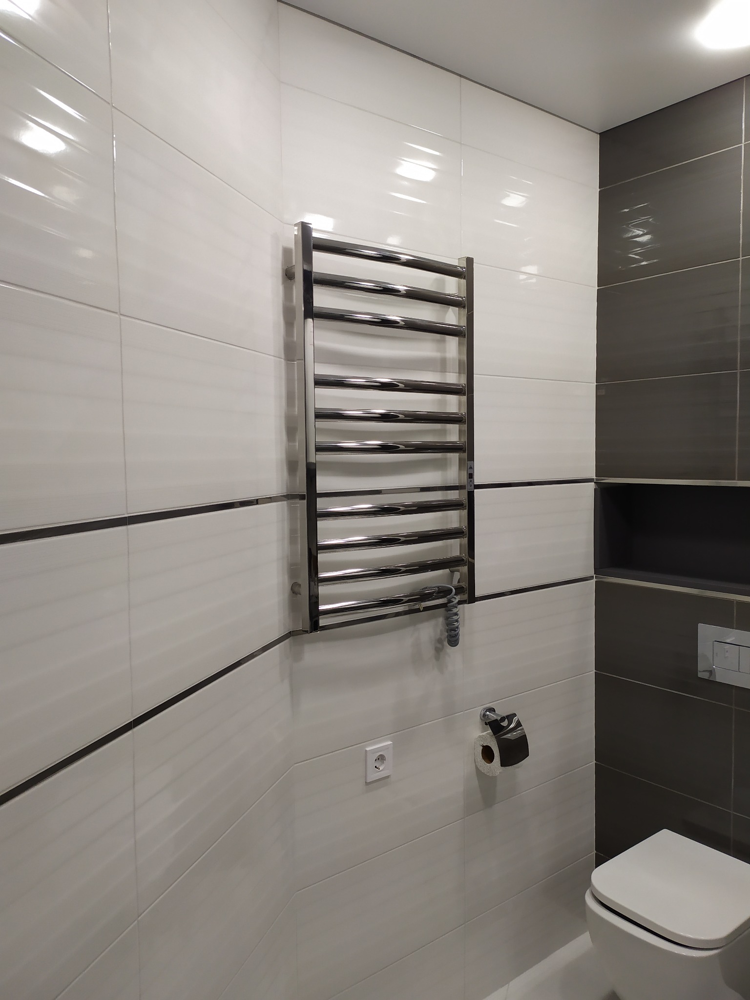 Nowoczesna łazienka z białymi i szarymi podłużnymi płytkami, chromowanym grzejnikiem drabinkowym, wiszącą toaletą i wnęką w szarej ścianie.