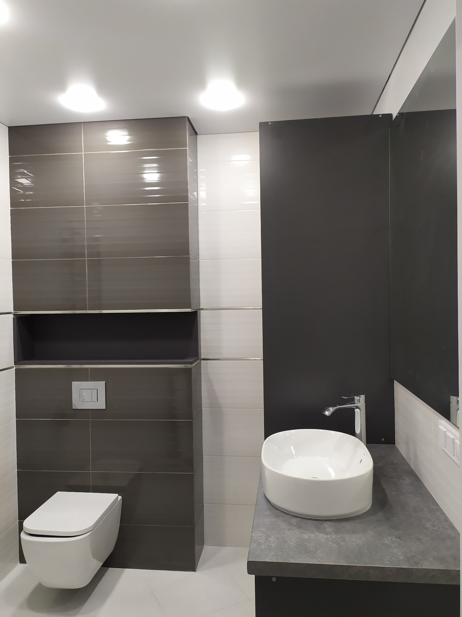 Nowoczesna łazienka z wiszącą toaletą, umywalką nablatową na szarym blacie i kombinacją białych i szarych płytek w połysku.