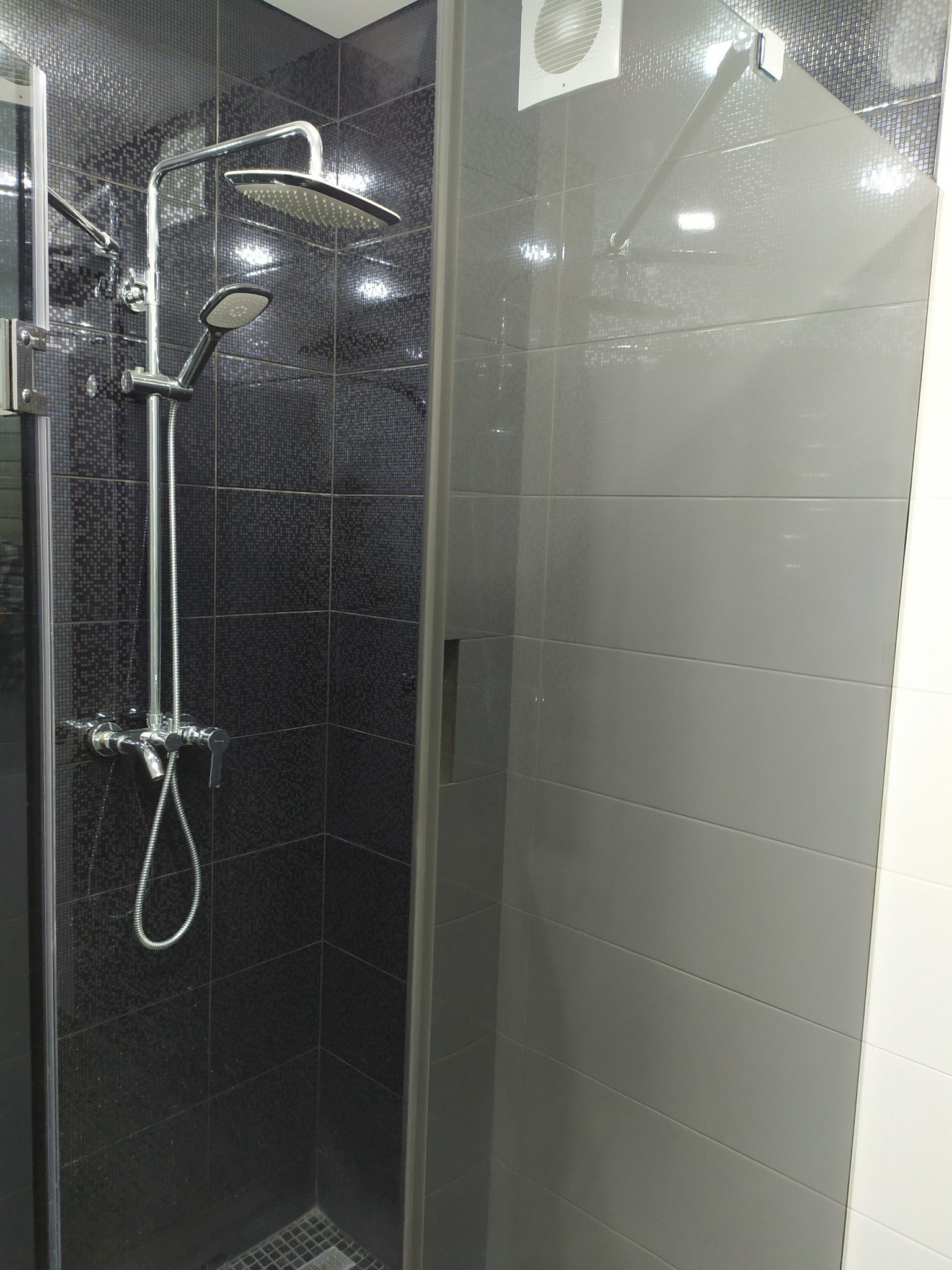 Nowoczesna kabina prysznicowa z deszczownicą i słuchawką, wyłożona czarną mozaiką i szarymi płytkami, z widocznym odpływem liniowym.