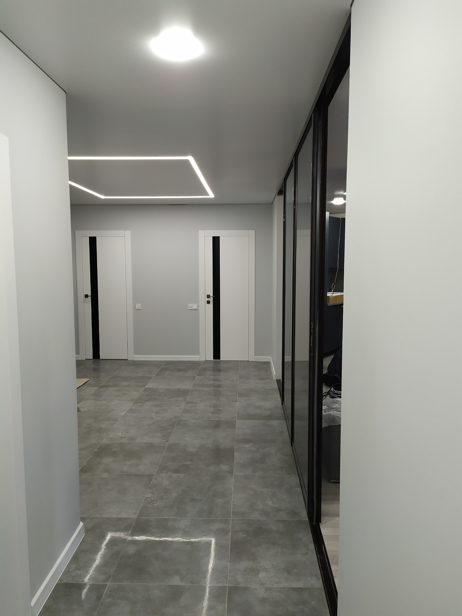 Nowoczesny korytarz z szarą podłogą z płytek, białymi ścianami, białymi drzwiami z czarnymi paskami i geometrycznym oświetleniem LED na suficie.