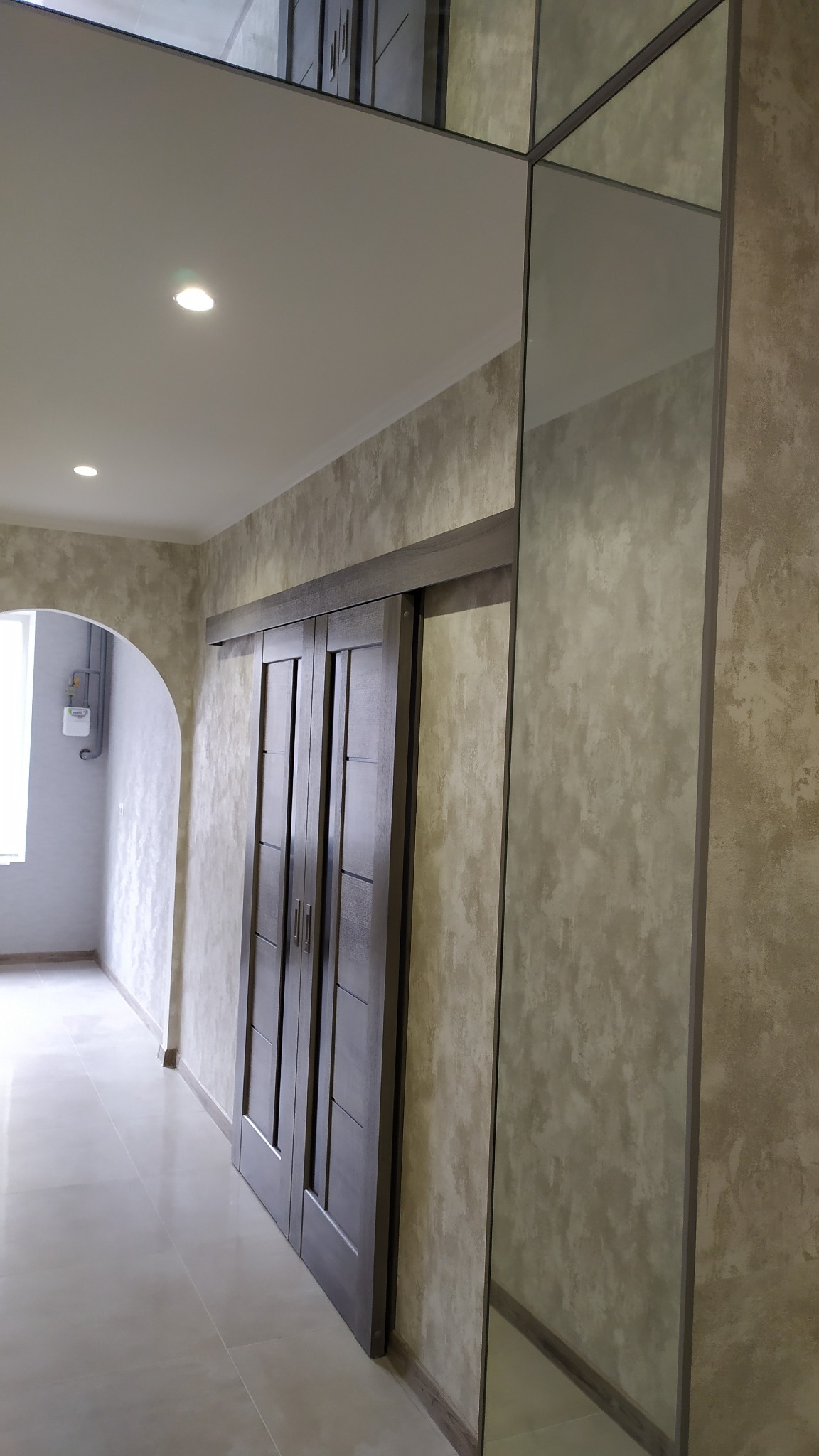 Wnętrze korytarza z przesuwnymi, ciemnymi drzwiami dwuskrzydłowymi i lustrzanymi panelami na ścianie, ściany wykończone w odcieniach beżu z efektem dekoracyjnym, jasna podłoga z płytek.