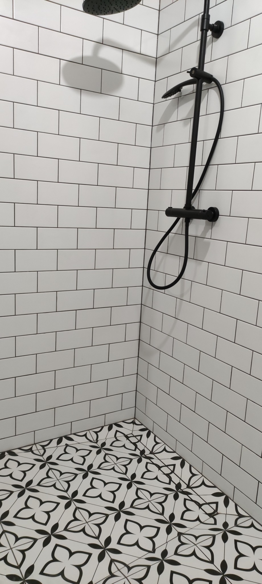 Wykończona kabina prysznicowa z czarną armaturą i biało-czarną wzorzystą podłogą, białe prostokątne płytki na ścianach z ciemną fugą.