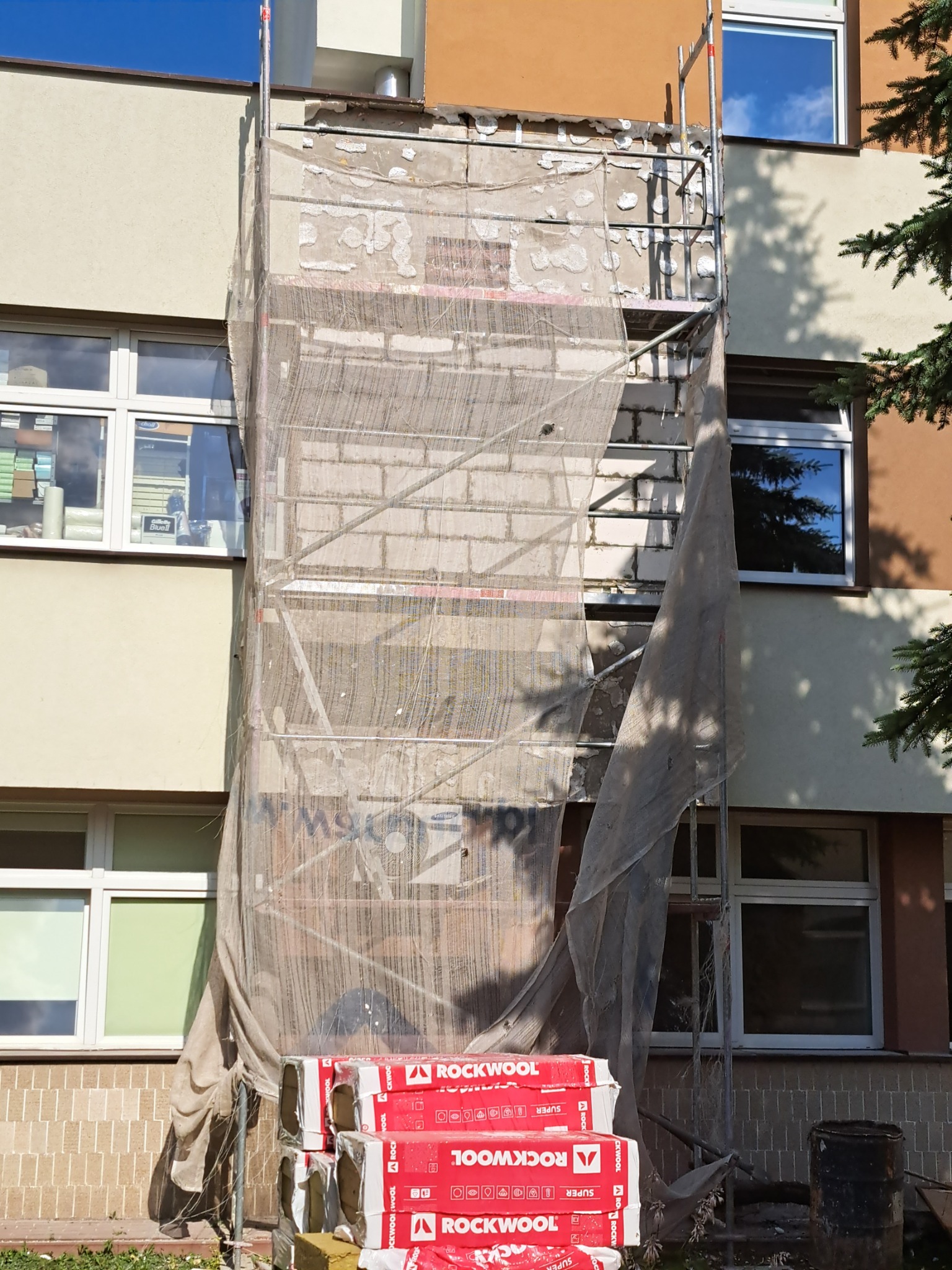Ocieplanie budynku: rusztowanie z siatką ochronną, odsłonięta cegła, paczki wełny mineralnej Rockwool u podstawy, fragment okna z odbiciem nieba.