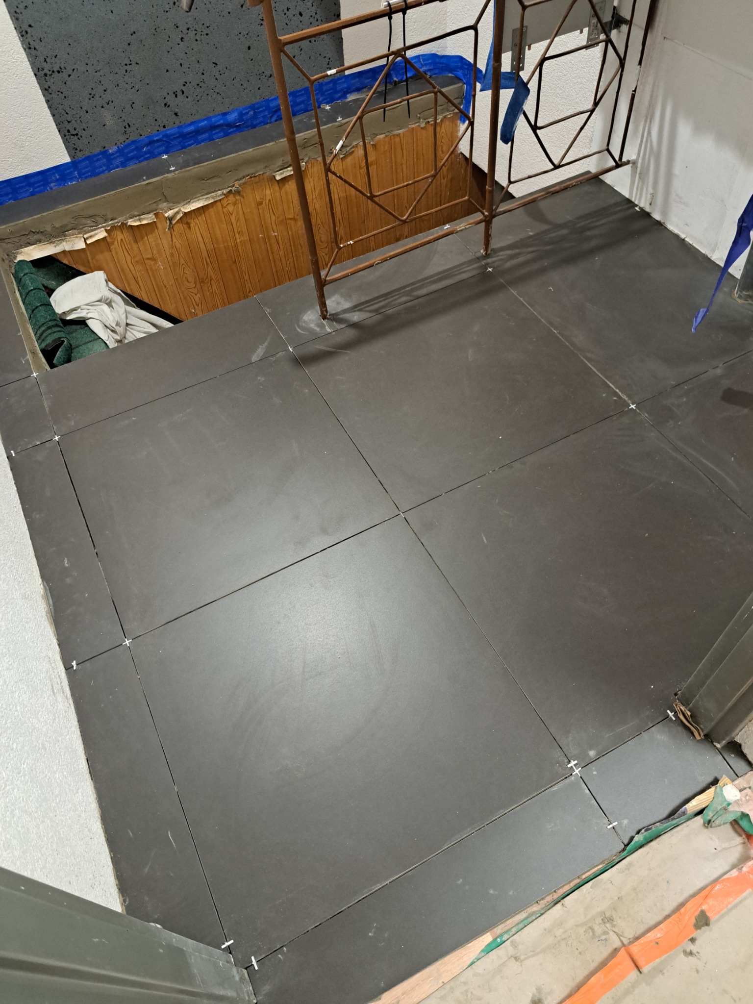 Ułożone ciemne płytki podłogowe z krzyżykami dystansowymi, widoczna krawędź z zabezpieczeniem i fragment metalowej barierki.