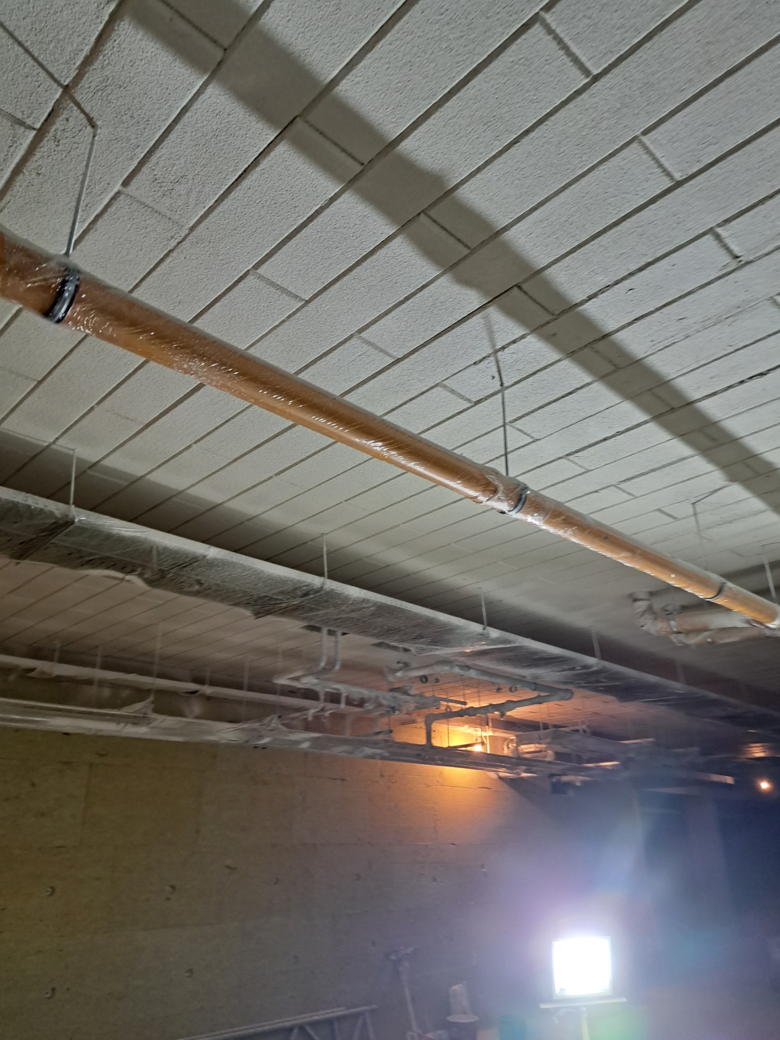 Ocieplony rurociąg w folii ochronnej, zawieszony pod betonowym stropem z widocznymi elementami instalacji i oświetleniem budowlanym.