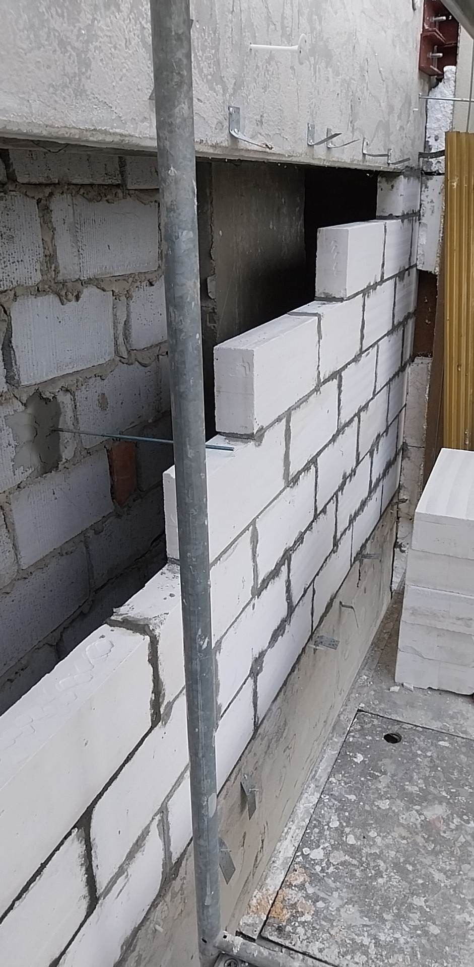 Wznoszenie ściany z białych bloczków betonowych, widoczne spoiny z zaprawy, fragment rusztowania i elementy mocujące do sufitu, surowy stan wykończenia.