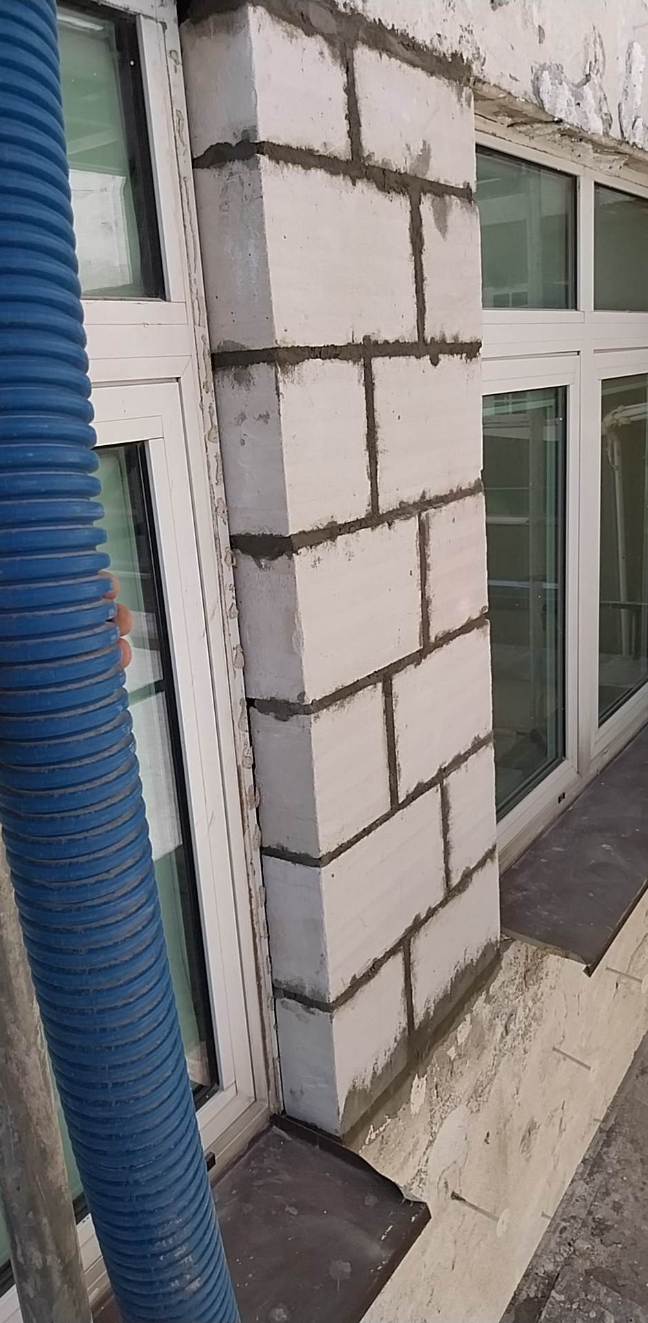 Wznoszenie muru z bloczków betonowych wokół okna, widoczny niebieski wąż wentylacyjny i fragment okna z szybami.
