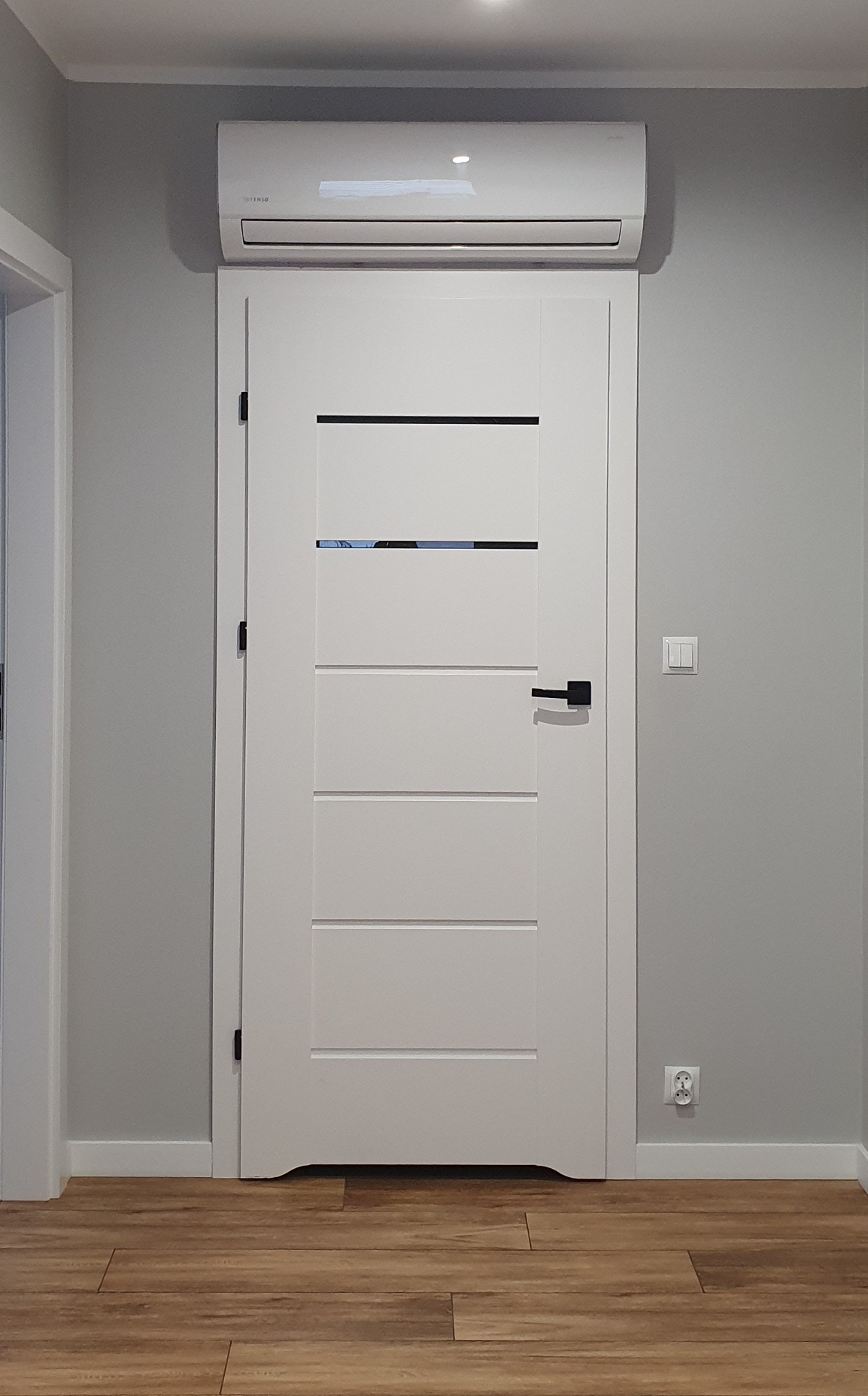 Biały klimatyzator zamontowany nad nowoczesnymi, białymi drzwiami z czarnymi akcentami w szarym pomieszczeniu z drewnianą podłogą.