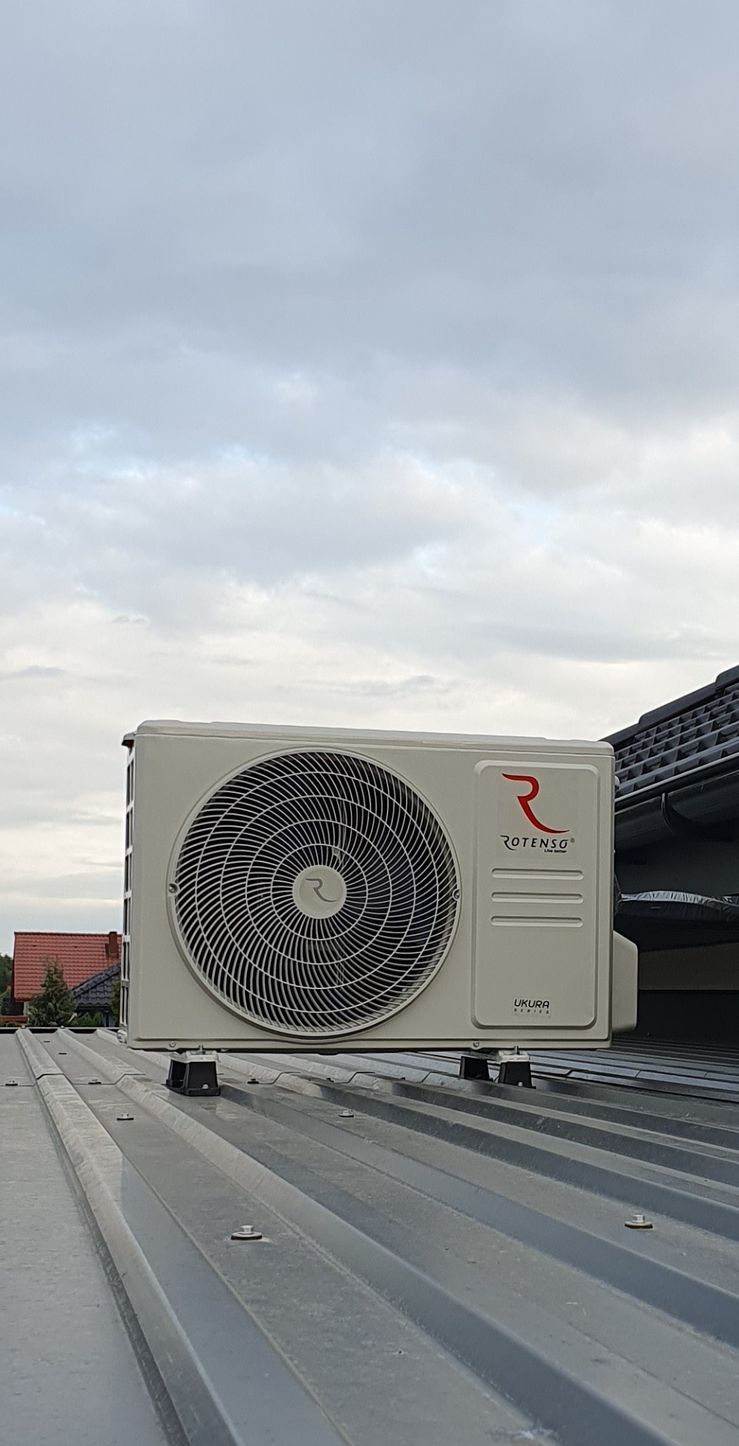 Zewnętrzna jednostka klimatyzacji Rotenso Ukura zamontowana na metalowym dachu z widocznymi śrubami mocującymi i pochmurnym niebem w tle.