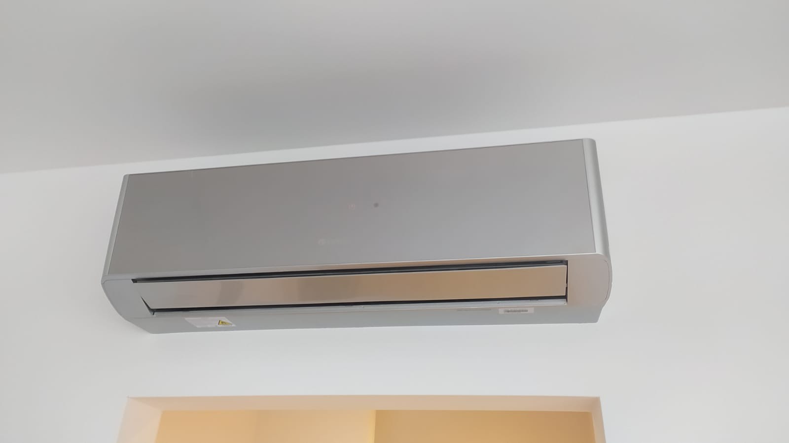 Nowoczesny, srebrny klimatyzator ścienny zamontowany w pomieszczeniu z białymi ścianami i beżowym fragmentem sufitu.