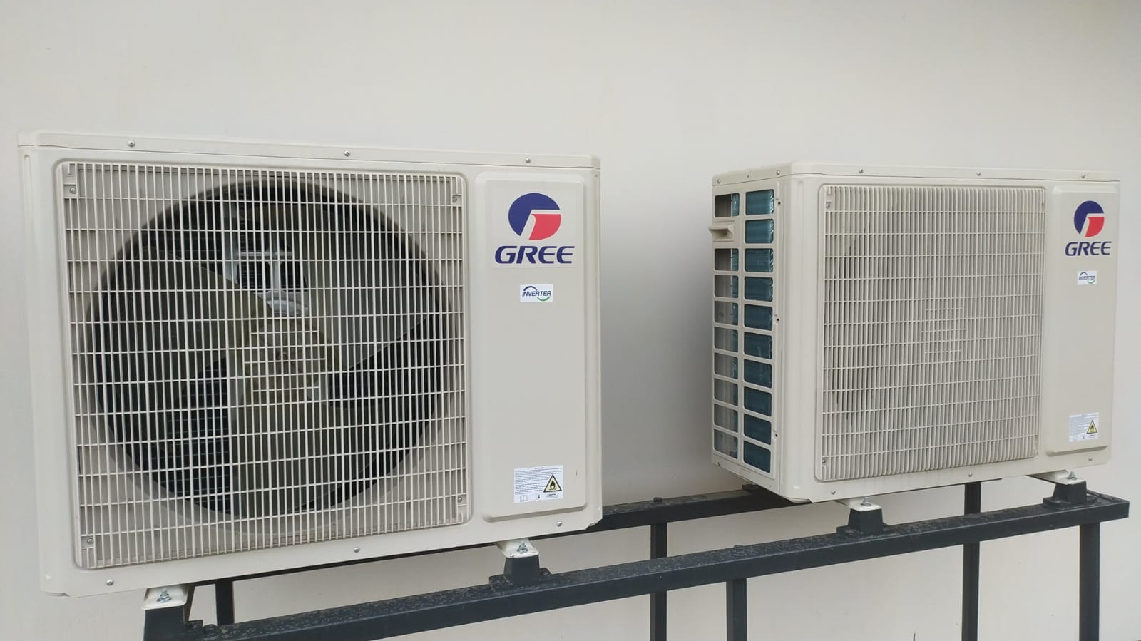 Dwa beżowe klimatyzatory marki GREE zamontowane na czarnej metalowej konstrukcji przy białej ścianie.