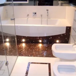 Nowoczesna łazienka z wanną obudowaną mozaiką, podświetlana podłoga z brązowej mozaiki, szklana kabina prysznicowa, biała ceramika sanitarna i półki z przezroczystego szkła.